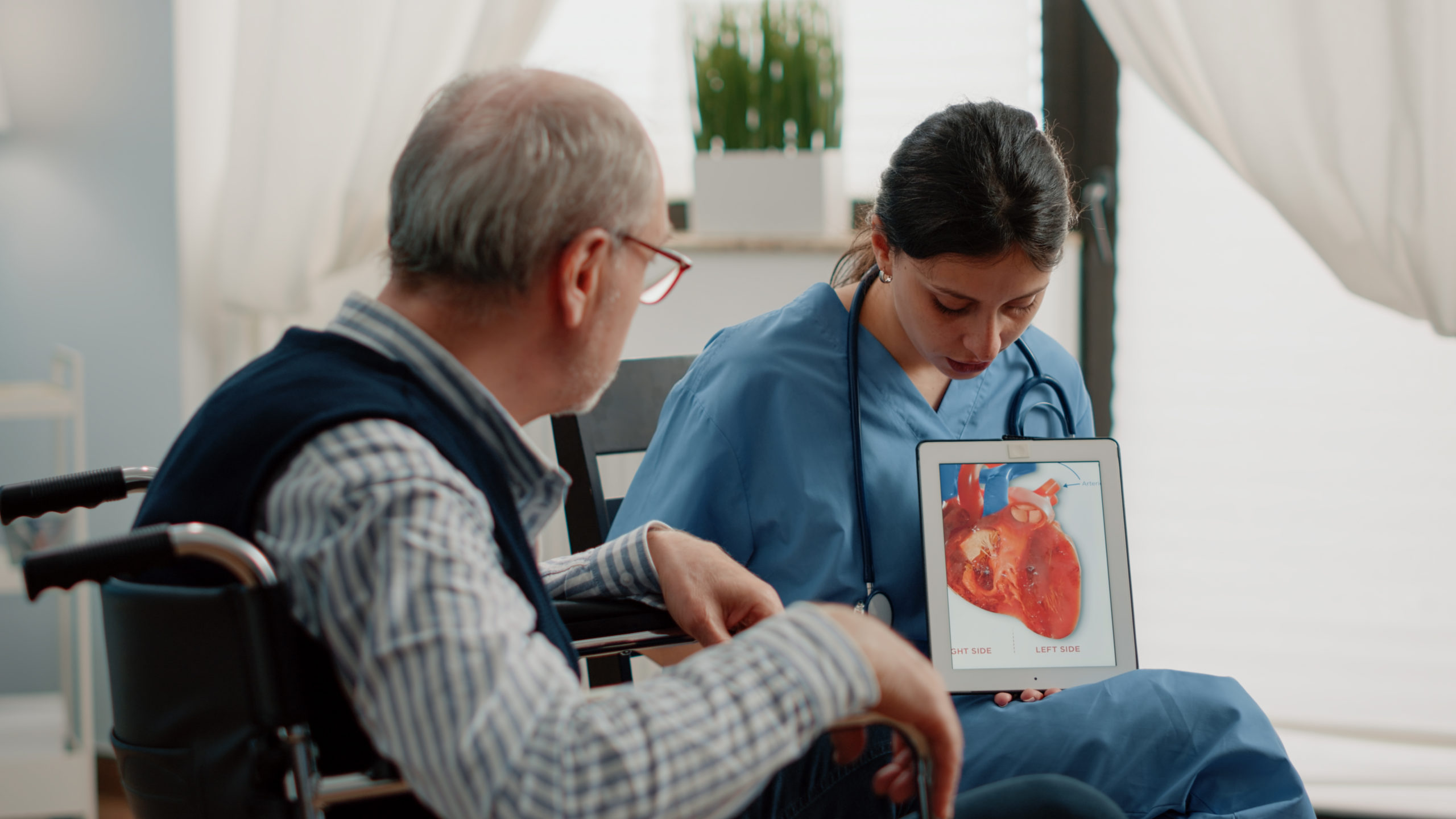 Nurse holding tablet with heart figure for cardiology diagnosis, showing cardiovascular issues to disabled patient at facility. Retired man sitting in wheelchair looking at blood vessel