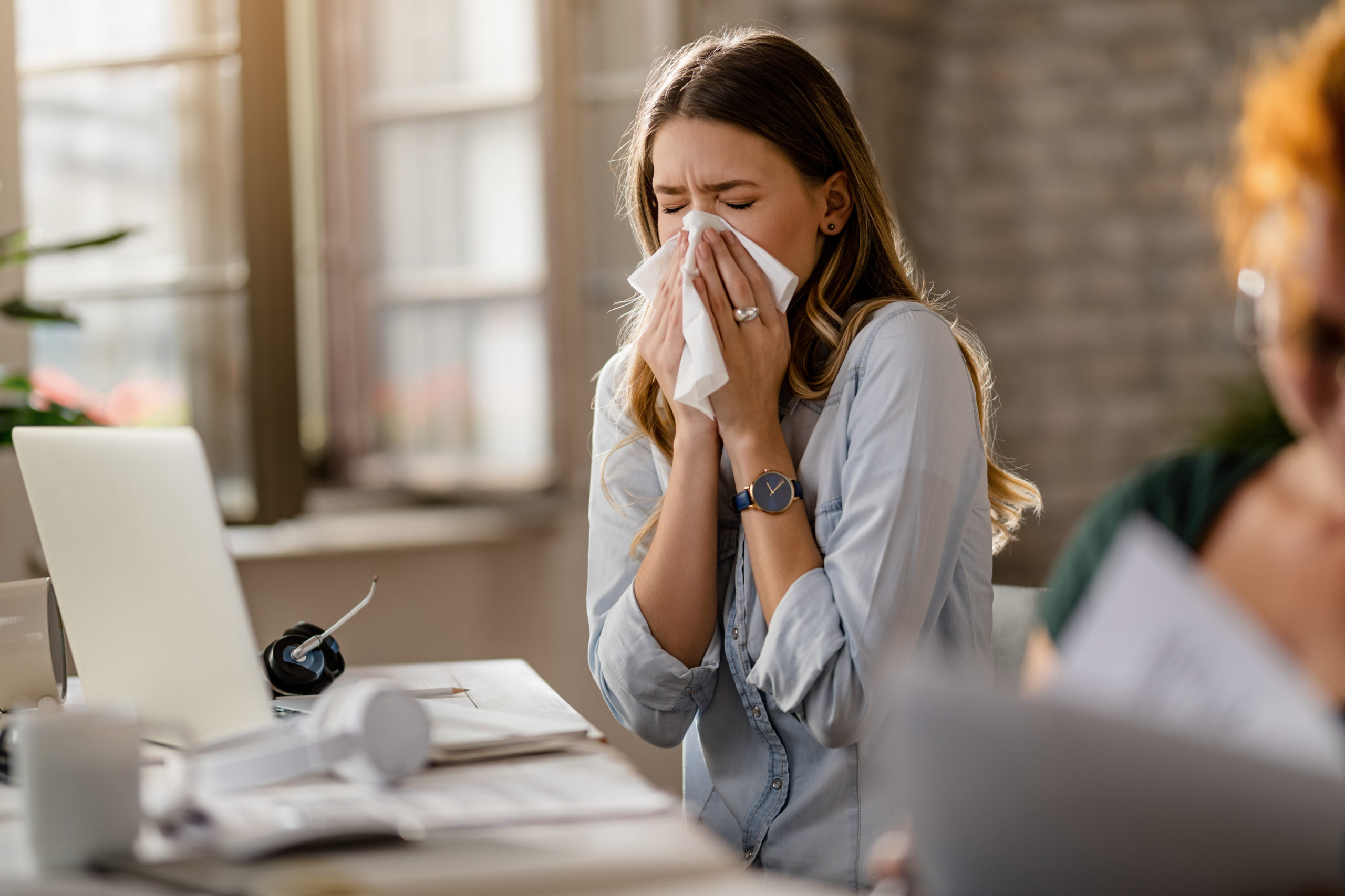 Young sick businesswoman sneezing in a tissue while working in the office.